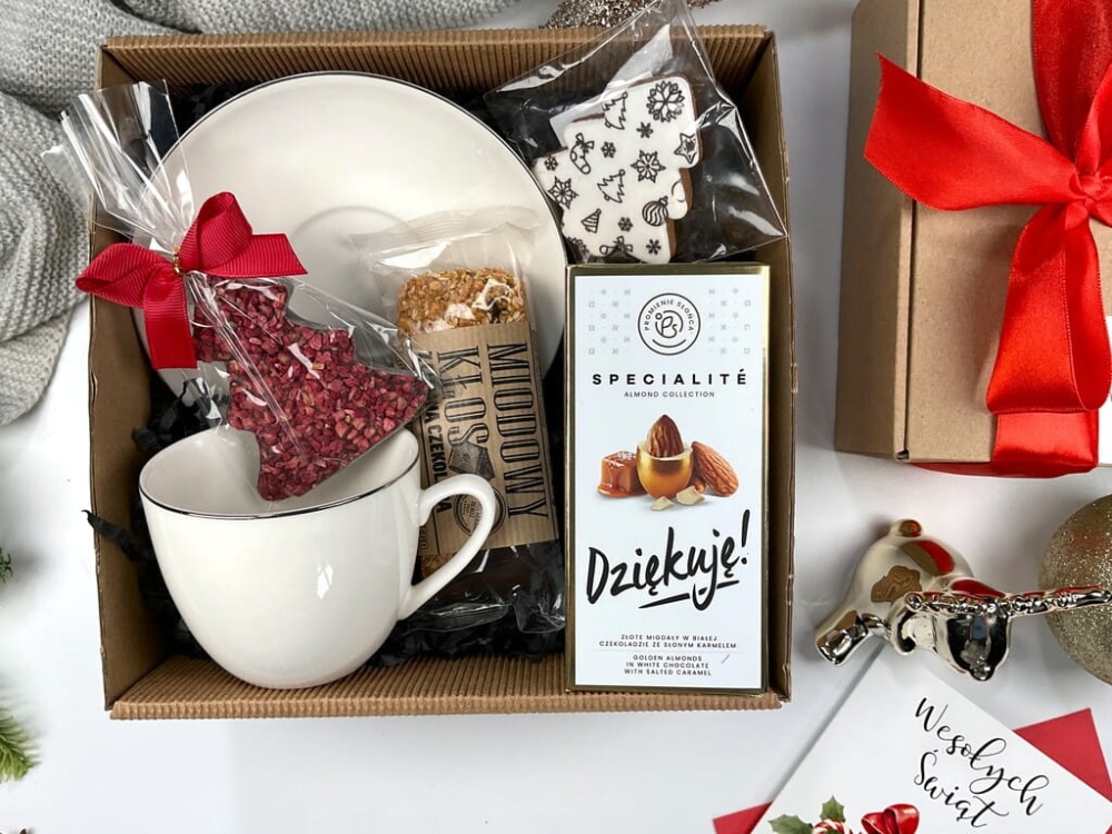 Świąteczny box prezentowy z białą porcelanową filiżanką ze srebrnym rantem, gorzką czekoladą choinka z malinami, batonem miodowym kłos z czekoladą, migdałami w złotej czekoladzie i ozdobnym pierniczkiem. Idealny prezent pod choinkę.