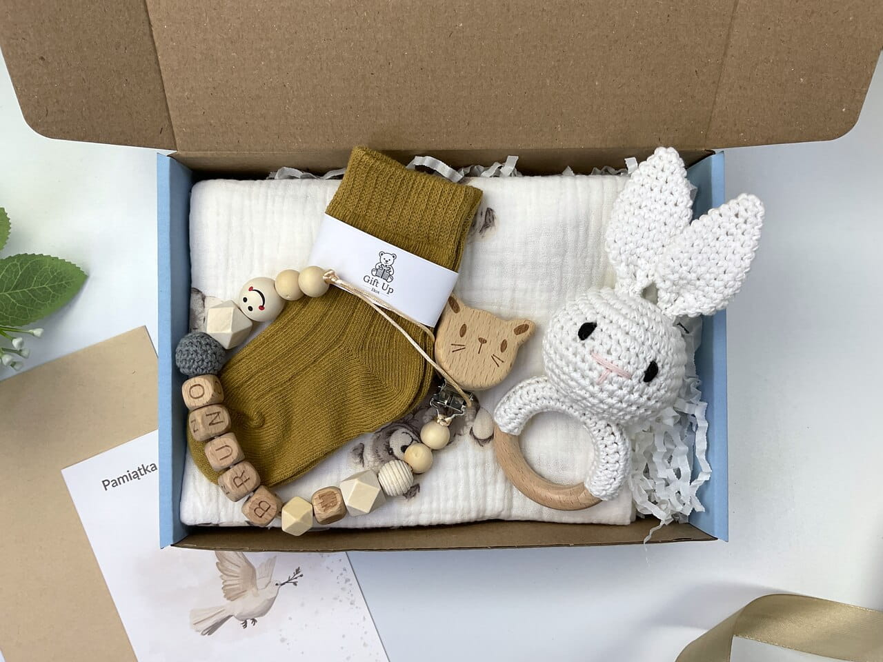 Zestaw prezentowy na chrzest dla chłopca: muślinowa pieluszka, ręcznie robiona grzechotka, zawieszka na smoczek, drewniana zawieszka z imieniem, skarpetki i personalizowana kartka.